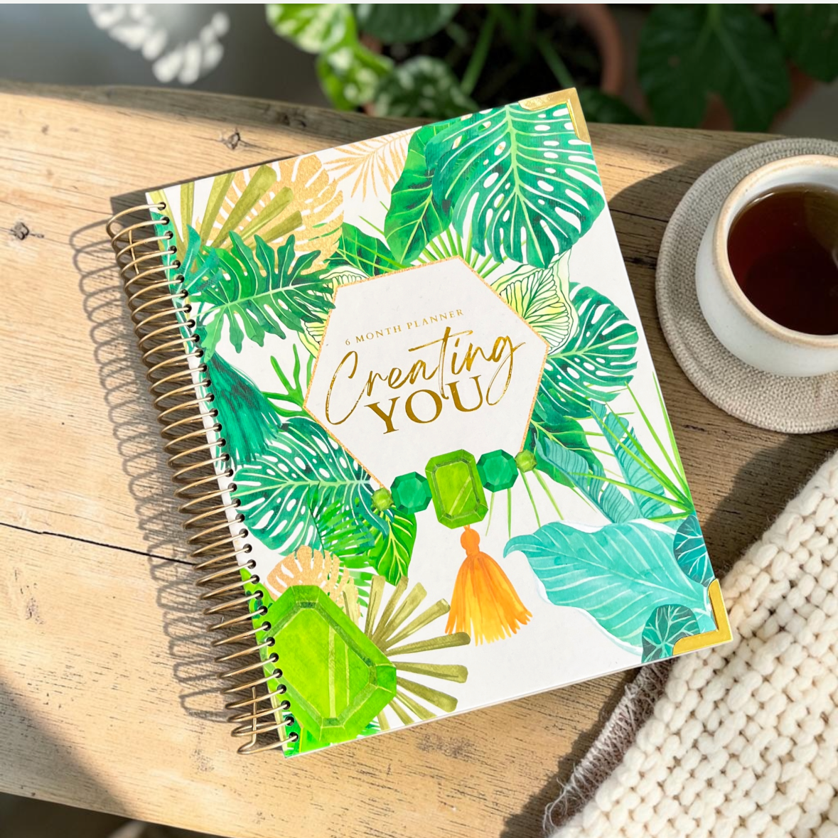 A spiral-bound notebook with a tropical leaf pattern and the words 'Creating YOU!' on the cover, shown resting on a wooden surface next to a cup of coffee. The image is displayed within a mobile app interface showing a page count of 4 of 4.