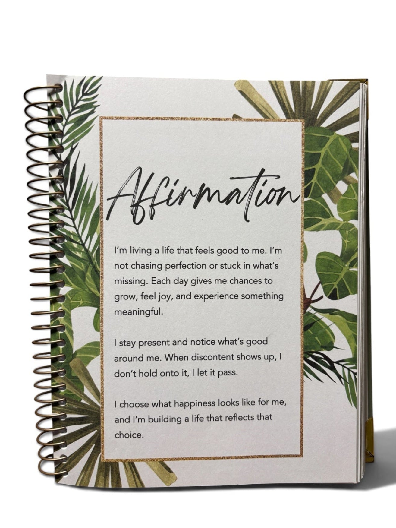 A spiral-bound affirmation journal with a white cover decorated with green tropical leaves and a gold border around the text, shown on a clean white background.