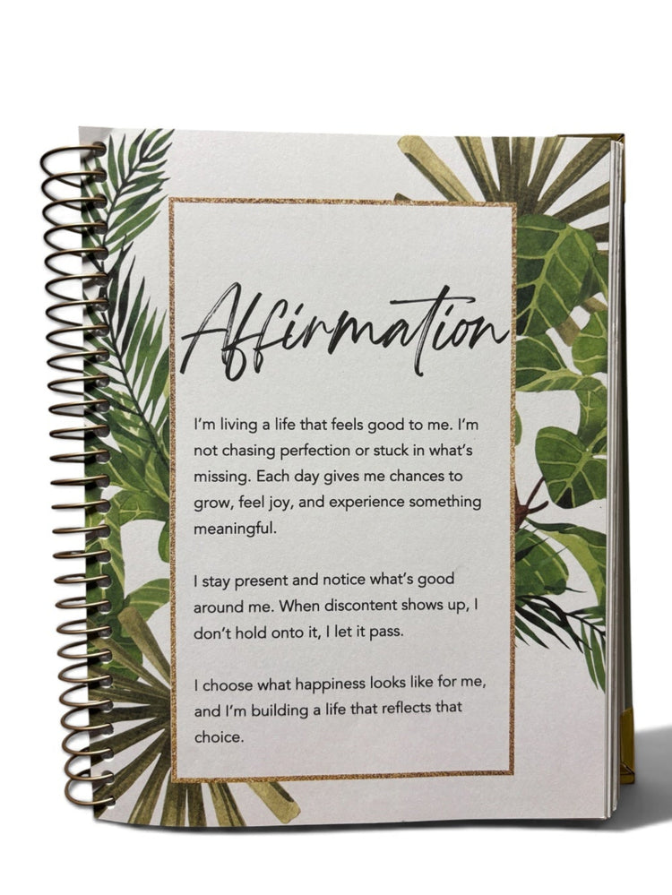 A spiral-bound affirmation journal with a white cover decorated with green tropical leaves and a gold border around the text, shown on a clean white background.