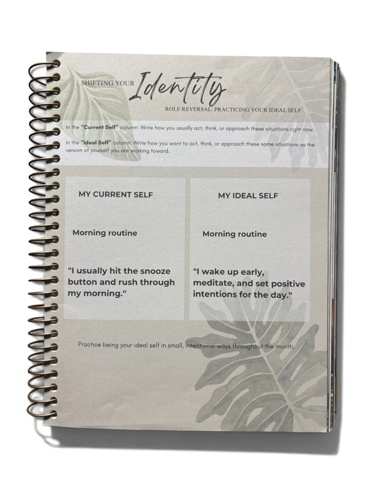 A spiral-bound notebook titled 'Identity' on a clean white background. The cover is a light gray-beige with black text and botanical leaf illustrations, showing sections for 'My Current Self' and 'My Ideal Self' with morning routine prompts.